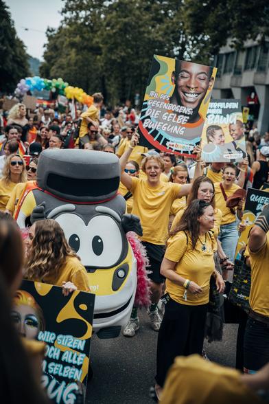 Das Maskottchen Bine bwegt zwischen Menschen in gelben T-Shirts bei der CSD-Demo in Stuttgart, auf einem bwegt-Plakat steht: "Teil des Regenbogens: Die gelbe BWegung"