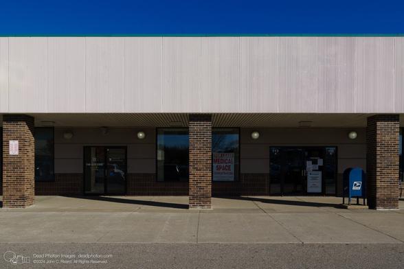 A photograph of a building facade of corrugated white-painted metal held up by thick brown-brick columns. Glass doors and windows are in shadow beneath an overhang, and signs read "Redwood Dental Group" and  "For Lease Medical Space." A blue mailbox sits next to one of the columns. A green strip marks the top of the wall, and there is a blue sky beyond.