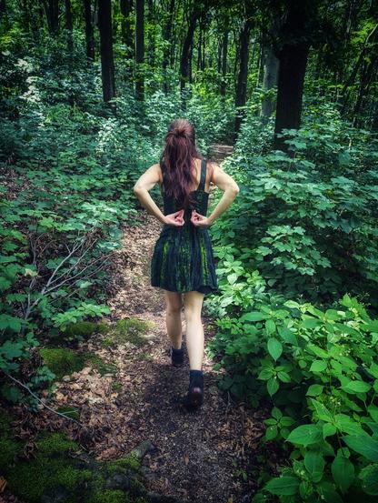 A woman in a dress of different greens walks through a forest of even more green tones. She reaches behind her back, to where a fairy would have wings.