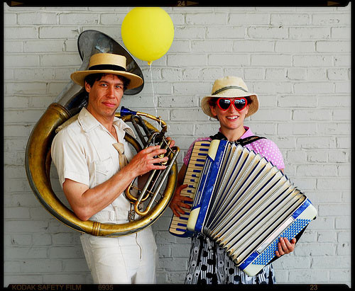 A man in a white suit wearing a tuba gazes unreadably at the camera, accompanying a piano accordion-wearing smiling woman in heart sunglasses.