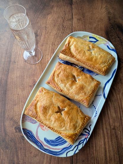 A rectangular photo of three golden-brown, rectangular puff-pastry slices sitting on an oblong plate, next to a champagne flute with a white-wine coloured drink in it. The plate is decorated with big blue flowers. They sit on a brown wooden background.