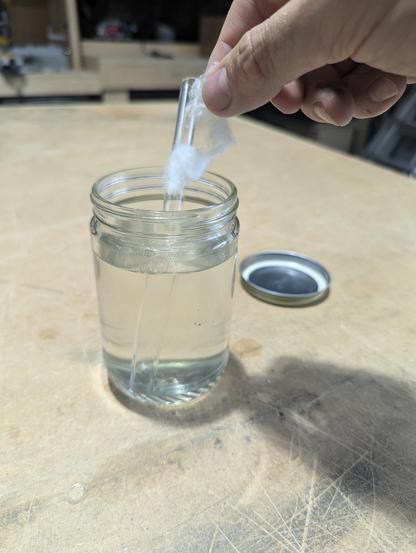 adding a pulled apart piece of the nitro cotton to a glass jar of solvent. There is a 12mm OD glass mixing tube in the jar that sits on a wooden table.