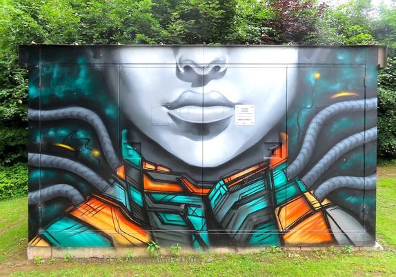 Street art on a large electrical box with the lower part of a face and a futuristic suit in green and orange with tubes attached; trees/hedges behind and grass around