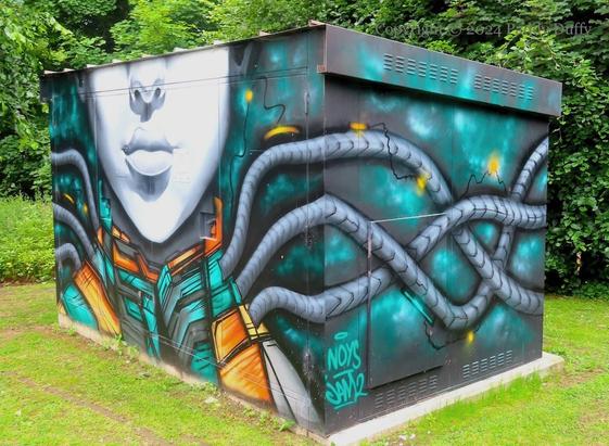 Street art on a large electrical box with the lower part of a face and a futuristic suit in green and orange with tubes attached; trees/hedges behind and grass around
