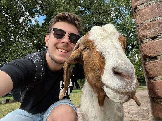 A young man wearing sunglasses and a black shirt is taking a selfie with a brown and white goat. They are outside with green trees and a blue sky in the background.
