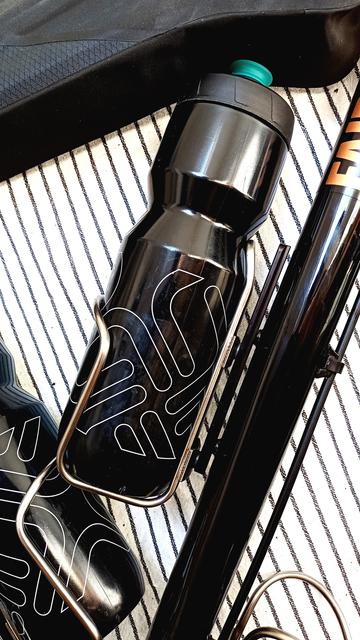 Ein schwarze Trinkflasche in einem silbernen Trinkflaschenhalter in einem Rahmendreieck. Am oberen Bildrand ist noch eine schwarze Rahmentasche zu sehen. Zwischen Rahmen und Trinkflaschenhalter ist ein Adapter zum passgenauen Positionieren des Trinkflaschenhalters montiert.