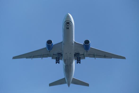 The photo shows a large passenger airplane flying in a clear blue sky with its landing gear clearly visible. The plane is in mid-air, with its landing gear extended. The underside of the aircraft is visible, including its wings, engines, and tail. The photo is taken from a ground perspective, looking directly up at the plane. (From Gemini AI)