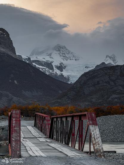 A wobbly looking one lane steel bridge with a patched up wooden deck across a river. In the background, an autumn forest, high mountains and a glacier