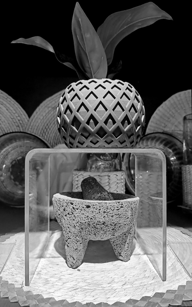 a black and white photo of a traditional mortar and pestle used in mexican cuisine at the front, a small flower pot on top and some plates and table mats in the background.