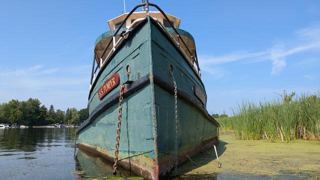 Bow-on view of a steam boat (?) on the Rideau river near Kars. The boat is derelict, resting in the muck on the river bank. Its green hull is covered with rust, and its hull plates are bowed in and puckered. The upper decks have been stripped of anything valuable. A name plate proudly proclaimed this the "Pumper".
