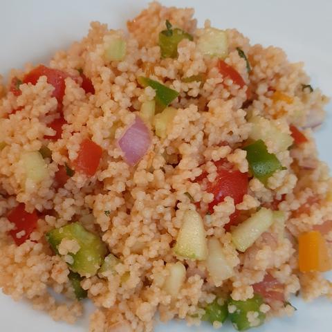 Photo of a serving of couscous salad on a white plate. Mixed in among the couscous are finely chopped tomatoes, cucumber, orange and green bell peppers, red onion, and parsley flakes. The tiny grains of couscous have a light orange color from the juices of the tomatoes.