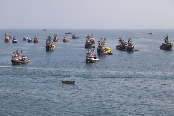 A bright but pale and hazy sky meets the horizon about a third of the way down. The sea that we're looking across is nearly flat, lightly rippled, and grey-blue. Across its surface there are over twenty fishing vessels, some of them very close together; all trawlers with different colouration on their hulls but generally all the same size and approximate shape with a very forward bridge and open deck space behind topped with cranes to manage nets, and almost all pointing towards us. Our viewing position is from above the height of the trawlers as we're on a larger ship heading out past the fishing boats.