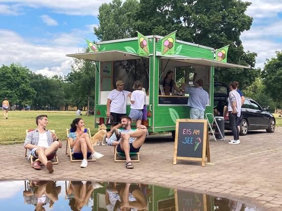 Ein Eiswagen in einem Park. Menschen auf Liegestühlen essen Eis und unterhalten sich.
