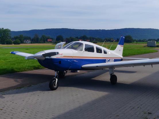 A blue-and-white low-wing light aircraft parked on the ramp seen from the front left. Grass, a few buildings and hills in the background. Thin red and gold stripes between the lower blue and the upper white part of the plane, the door on the right side is open. Type is a Piper Archer III, registration, partly obscured by the wing, is SE-MMP.