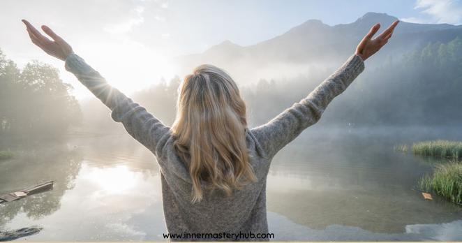 A person with long hair stands by a misty lake, arms raised towards the sky, facing the mountains in the distance.