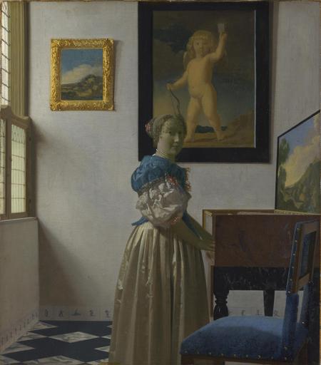 Vermeer's painting Woman Standing At A Virginal showing a woman in a long cream dress with a blue bodice standing at an old fashioned Virginal with two paintings on the wall.