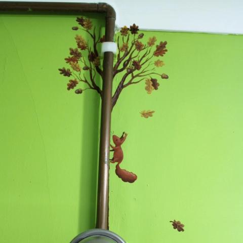 Green wall with a copper pipe. Behind the pipe there is a painted oak tree with a red squirrel climbing up it.