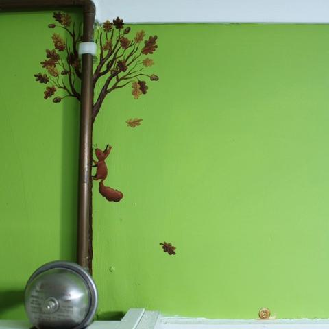 Green wall with a copper pipe. Behind the pipe there is a painted oak tree with a red squirrel climbing up it. There is a small snail above the nearby doorframe.