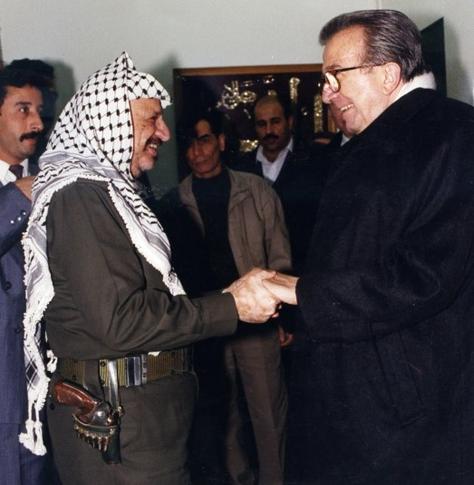 Arafat e Andreotti si incontrano all'inizio degli anni '80. Si stringono la mano. Sullo sfondo le guardie del corpo. Intorno alla cinta di Arafat, vestito in uniforme militare e con la kefiah, si nota una rivoltella.