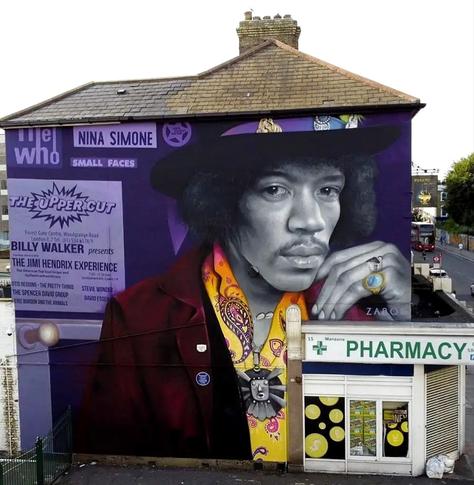 Streetartwall. In memory of the legendary concerts at "The Upper Cut" club in London, a fantastic mural featuring Jimi Hendrix was sprayed/painted on the outside  of a two-story  building (12 x10 m). The image is photorealistic and shows him in a purple suit and an orange paisley shirt. He looks at the viewer with a penetrating but calm gaze. His hand is casually resting on his face. Below the mural is a pharmacy. He is practically leaning on it. Next to Jimi is a purple text that refers to the club and looks like an old advertisement. It reads: "The Upper Cut: "Billy Walker presents
The Jimi Hendrix Experience"
as well as advertisements for stars such as Nina Simone, The Who, Small Faces, The Animals
Stevie Wonder and many more.
Info: The Upper Cut Club was a former ice skating rink that owner Billy Walker converted into a "plush big beat palace" for 200,000 pounds. The concert list in the opening week of 1966 was breathtaking. All the stars were represented. Incidentally, Jimi Hendrix's first appearance at the Upper Cut cost just five shillings admission.