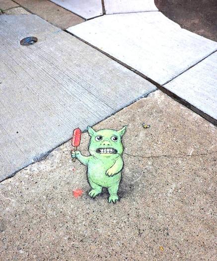 Streetart. On a gray sidewalk with four small holes in the ground, a small green monster (named Maurice) was drawn on the ground with chalk. The four holes have been integrated into the drawing and are now the eyes of the green friend. He is standing there looking scared with his mouth open and holding a red popsicle in his hand. A bee flies next to him. The ice cream is already dripping down. Title: "Maurice is caught between his two greatest fears: bees and brain freeze."