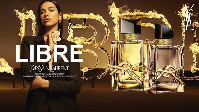 Dua Lipa models for YSL Libre Flowers & Flames fragrance with a Moroccan desert setting; the word Libre appears to be in flames behind Lipa, who is wearing a black outfit. The fragrance bottles are shown on the right half of the image.