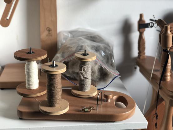 Three wooden bobbins sit on a tensioned lazy kate (spool holder) with small samples of handspun wool yarn - cream cashmere, brown camel, and grey yak. An open ziplock bag full of yak fiber sits at the back of the white chest of drawers behind the yarn on the base of an Ashford yarn swift, next to the spinning wheel on the right.