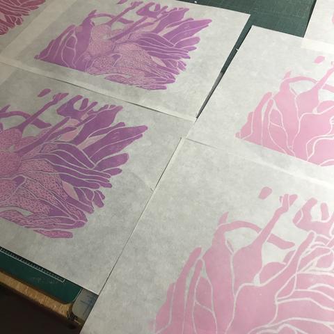 A collection of at least five 9.29” x 12.5” prints drying on a tabletop. Half show my print in its first state with bulbous pale rose bubble tip anemones. Half show the second state with the anemone over printed with texture in purple pink.