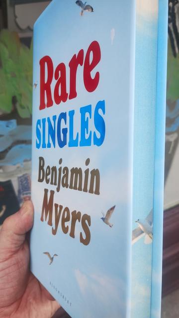 Rare Singles by Benjamin Myers, light, sky blue cover with clouds and birds, held side on to show the spredges (painted edges) depicting more sky and a bird