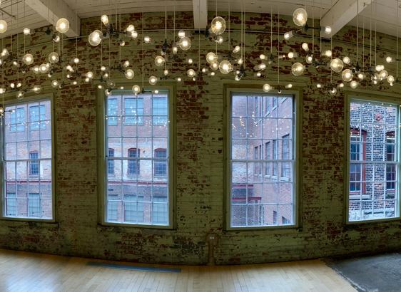 Panoramic distortion of art display with white lights of varying sizes descending from ceiling before a wall with rough brick and four latticed windows through which other buildings are visible.