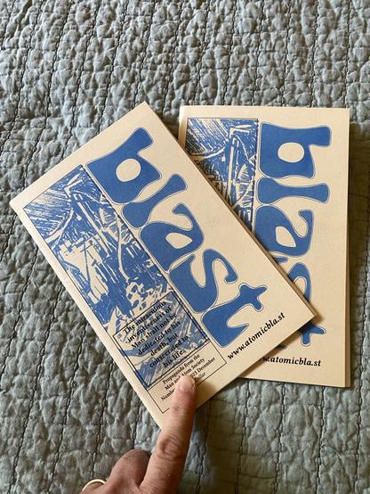 Two copies of Blast magazine/fanzine. Finger holds cover in place. Bedspread in background.