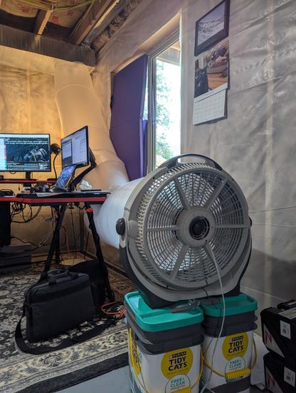 A very, very, very jank tube of painter's plastic funneling air from a circular floor fan into HVAC ductwork on the ceiling. The tube has wedged itself between a messy desk with two large computer monitors, and the unfinished wall of the basement. Image backlit by a window half-covered with a folded yoga mat.
