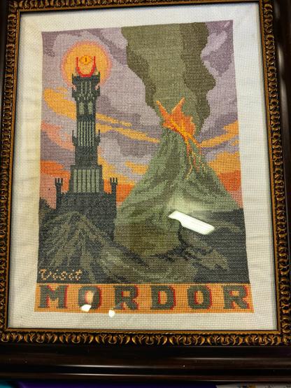 Framed cross-stitch artwork depicting the dark tower and Mount Doom from "The Lord of the Rings," with "Visit Mordor" stitched at the bottom.