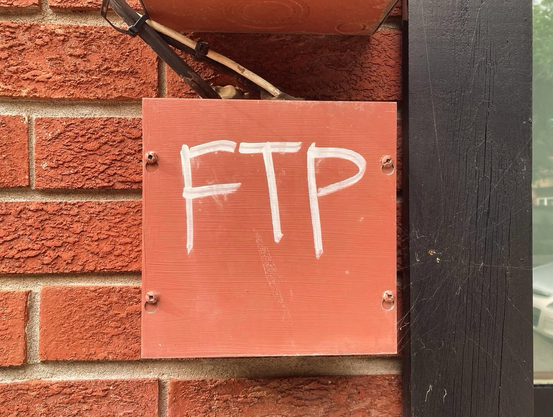 The letters “FTP” painted in white letters on a small, red utility box on the outside of a red brick building
