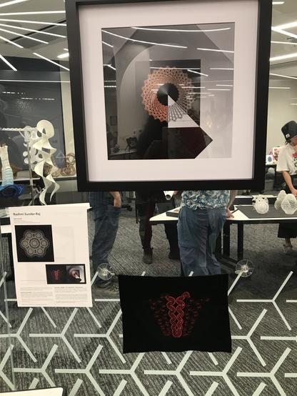 A predominantly black, grey, white and red square print in a black frame with a white mat on a glass wall. There is a squiggly piece of crochet using red sequins and red and black metallic thread on a black background below it. There is an information sheet off to the left.