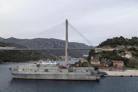 An overcast day and we're on a ship sailing out of a port, passing a channel between two land masses that are connected by a suspension bridge. Docked alongside the nearest of the land masses on the right of us is a boxy-looking naval vessel in typically grey paintwork with a textured look across it all like corrugated iron. A low bridge sits towards the front, barely jutting over the bulky shape of the ship from this angle. At its rear is a flat area of deck with a flight deck on which to land helicopters. It all looks functional and industrial rather than military.