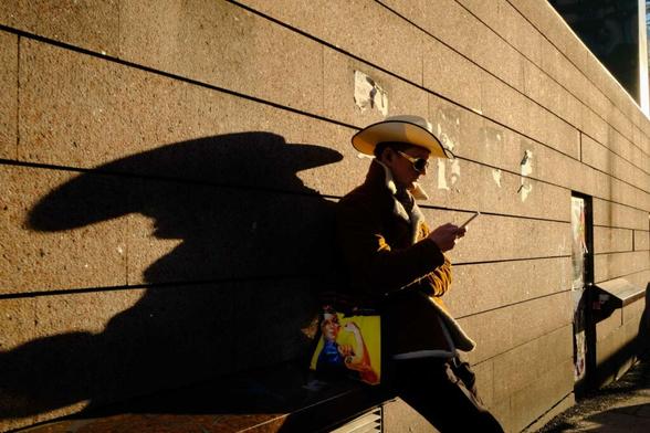 A man in a cowboy hat browsing on his phone. He is carrying a tote bag with feministic symbolism on it. The sun is low, and he cast a long shadow behind him.
