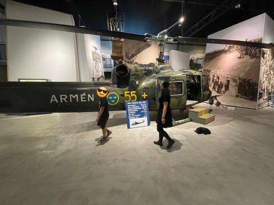 Jen and a kid is walking past a swedish military chopper inside a hangar