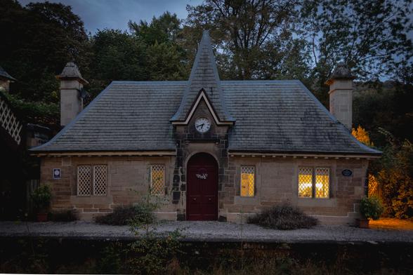 The old Victorian stationbuilding just before darkness. Frontage with a door in the middle; a white clockface in the gable; two windows on either side.  Artificial light is glowing from the windows on the right.