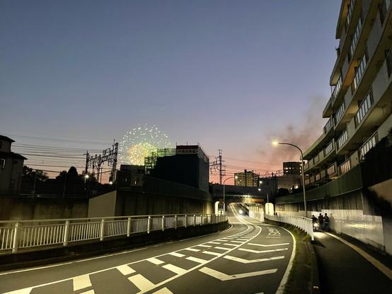 Looking along a curving empty street after sundown, the sky is still bluish and pink on the horizon. The street curves off into the distance as it passes under a train overpass and heads up on the other side.

In the centre of the frame is the silhouette of a building under construction, and just peeking above one corner of the building are several bursts of fireworks, mostly green with some red.

Several stories of an apartment building frames the right side of the image, the building is curved to match the street, the glass on the balcony railings reflect the last pink and blue light from the sky.