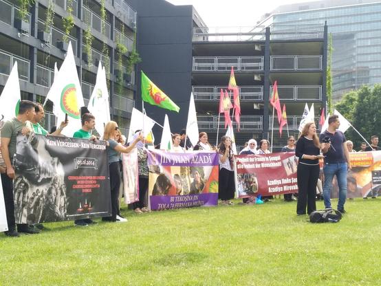 Wiese vor dem Landtag mit einer geschlossenen Reihe aus Menschen mit Transparenten und Fahnen der Jesiden. Davor Wissler am Mikrofon.