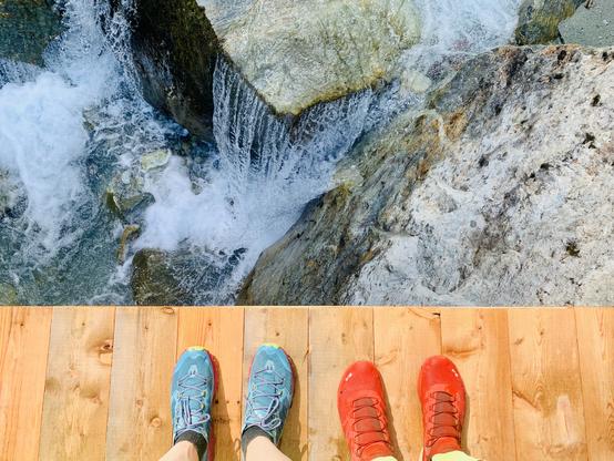 Two person standing on a wooden bridge. Just feet are visible. The water below flows over large stones.