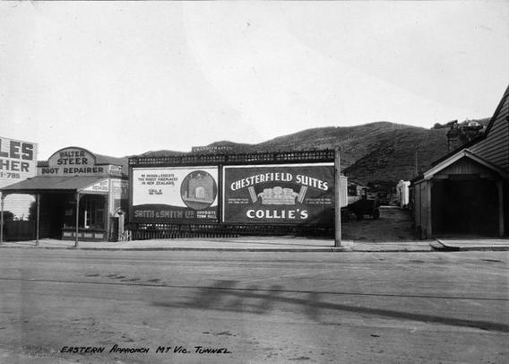 Black-and-white photo: Billboards beside the eastern approach to the Mount Victoria Tunnel, 'Chesterfield Suites' and 'Smith & Smith' for fireplaces. 1929. Photographer, F. G. Barker. Description: A view across a sealed road and footpath to a small boot-repair shop at left, two billboards in the centre and to the right an unsealed driveway or narrow lane. Hills are in the background. Citation: Wellington City Council Archives, 00127-1-Page 41. https://archivesonline.wcc.govt.nz/nodes/view/139090