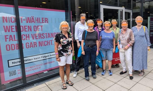 Gruppenbild von 8 Frauen. 3 Frauen im Hintergrund und 4 Frauen vorne.
Sie stehen vor einem Gebäude mit einer großen Glasfront. Der Platz auf dem sie stehen hat Bodenfliesen.
Hinter dem Fenster ist ein Überlebensgroßes Plakat auf dem steht: "Nicht Wähler werden oft falsch verstanden oder gar nicht".