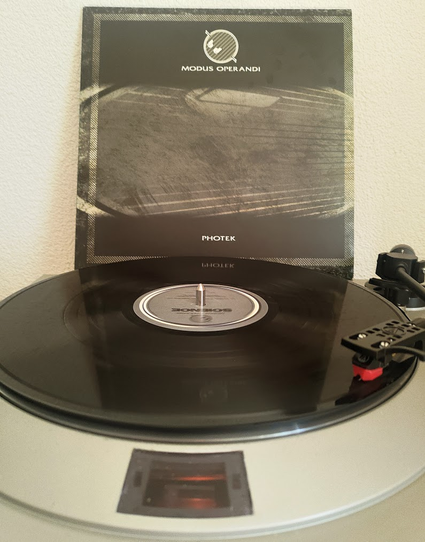 Photek's Modus Operandi playing on a Denon DP-30L turntable. The cover in the background is monochrome black/grey/white. At the top is the Photek Logo and the title of the record, and at the bottom the artist's name.