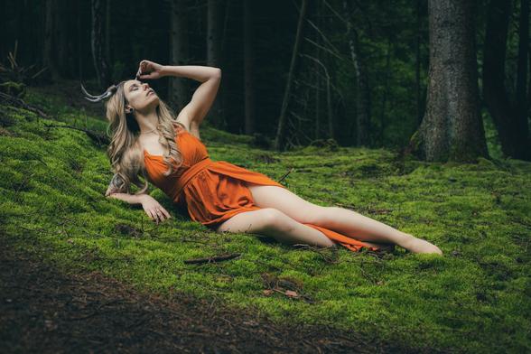 "A woman with long blonde hair and a tattoo, wearing an orange dress, is reclining on a lush, moss-covered forest floor. She is gazing upwards with her hand on her forehead, surrounded by tall trees and a serene, green environment."