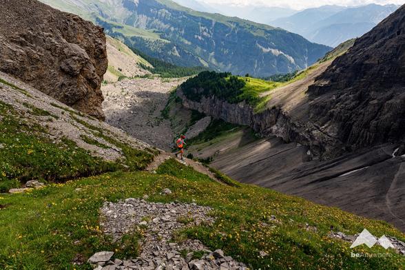 Trail runner in an orange shirt running down a hiking trail in a fascinating mountain landscape. Yellow flowers on the green meadows. beAnywhere logo in the lower right corner.