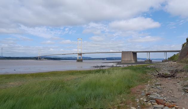 Old Severn Bridge, closer view.  A suspension bridge across the Severn Estuary.  There is grass in the foreground.  Aust cliff rises to the left.  The Welsh coast is visible at the far side.