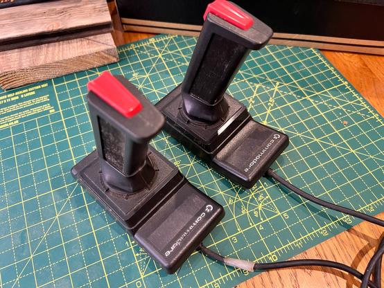 A photo of two dark grey Commodore branded joysticks. The handles are very straight, leaning forward, with a single red button on the top. They were never great to use.