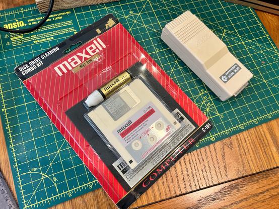A photo of a Maxell branded set of floppy head cleaners for both 3 1/2” and 5 1/4” drives plus some cleaning fluid, which I imagine would be useless now. Next to it is a TV modulator for the Amiga 500, a long beige box.
